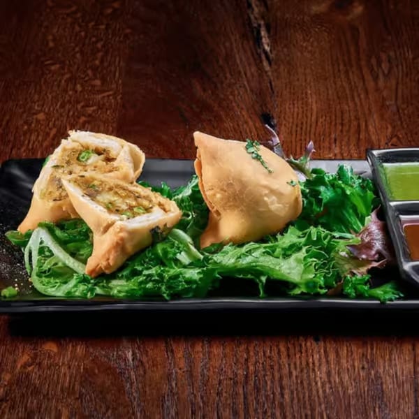 Crispy samosas, one cut open, served on a bed of lettuce with two dipping sauces.