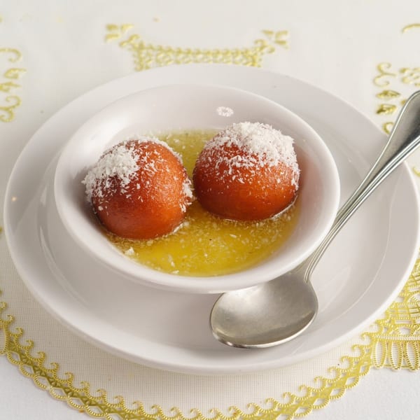 Two gulab jamun, topped with shredded coconut, in a white bowl with syrup and a spoon.