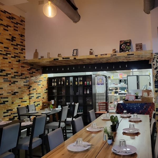 Interior of a restaurant with brick walls, wooden tables, and set place settings, with a kitchen visible.