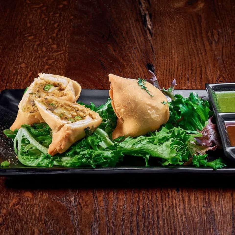 Crispy samosas, one cut open to show the filling, served on a bed of lettuce with dipping sauces.