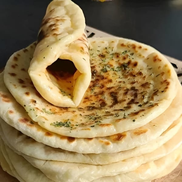 A stack of freshly baked naan bread, with one piece rolled up and seasoned with herbs.